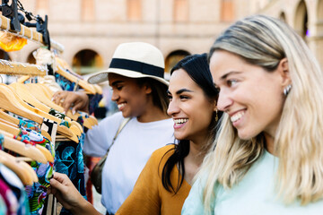 Three young happy women looking at clothes at weekly clothing market. Female tourist having fun enjoying shopping day looking at second hand clothes at town outdoor market