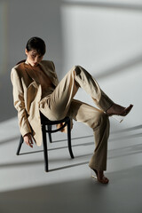 A woman in a beige suit sits on a chair against a white backdrop.