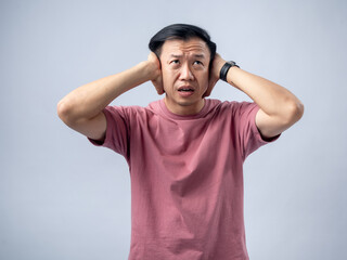 An Asian man in a pink t-shirt holds his head in both hands with a distressed expression, eyes...