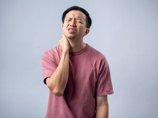 A man in a pink shirt holds his neck with a pained expression, indicating discomfort or neck pain. The plain light blue background emphasizes his distress and highlights