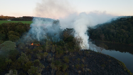 Smoke in the air near the lake and vegetation. Forest fire in the dry season in the rural area of ​​Apucarana with trees being burned.