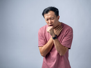 A man in a pink shirt coughs violently, holding his throat with one hand and covering his mouth with the other. His expression shows discomfort and pain. The plain light blue background