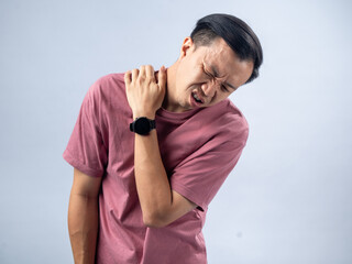 A man in a pink shirt holds his shoulder with a pained expression, indicating shoulder pain or discomfort. The plain light blue background emphasizes his distress