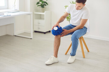 Portrait of a young man applying cold compress to leg sitting on chair at home. Male person holding...
