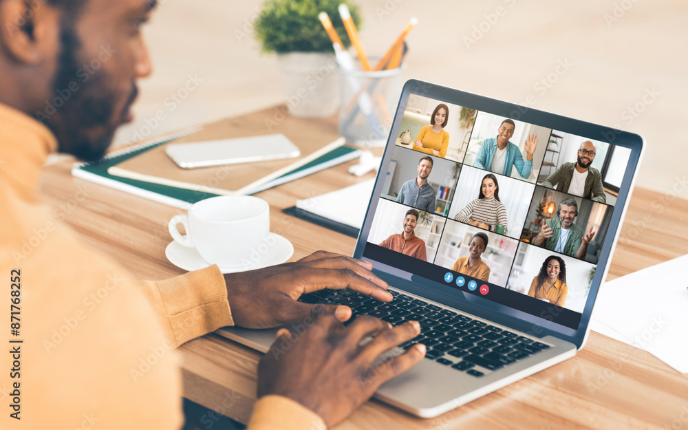 Poster black man sits at a desk in front of a laptop, participating in a virtual meeting with a group of pe