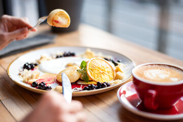 Top view of a woman eating syrniki — cottage cheese fritters with strawberry jam. Female hands holding fork and knife over homemade breakfast pancakes.	