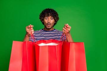 Portrait photo of young indian curly funny shopaholic man student in striped t shirt shocked looking into his bags isolated on green color background