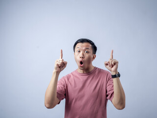 Asian man in a pink t-shirt, standing against a plain background, pointing upwards with both index...
