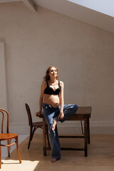 Beautiful pregnant woman in black top and jeans barefoot, sitting. Big pregnant naked belly. Pretty woman waiting child. Sofa, small window, white walls