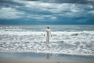 Hairless girl with alopecia in white futuristic suit comes out of cold restless sea on sandy beach,...
