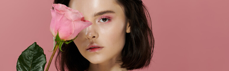 Obraz premium A woman with dark hair poses in front of a pink backdrop, a single pink rose obscuring one eye.