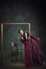 Obraz premium Portrait of curious elegant young woman, royal person peek out picture frame with interest against dark green background. Mystery of future. Concept of history, comparison of eras, remake