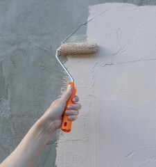 A hand holds a paint roller and applies beige paint to a previously grey wall, creating a stark...