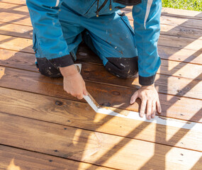 A worker in blue overalls applies painters tape to wooden deck boards in preparation for a painting...