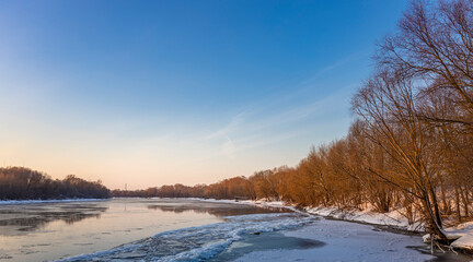 A winter sunset casts a warm glow over a frozen river, the icy water reflecting the sky. Bare trees stand along the banks, their branches reaching towards the clear blue sky..