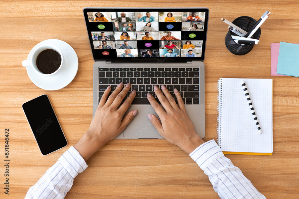 Poster A person is sitting at a wooden desk and using a laptop to participate in a video conference. The laptops screen shows a grid of video tiles, each displaying a different participant, top view