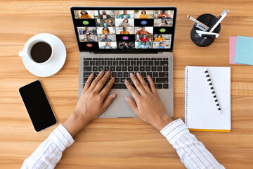 A person is sitting at a wooden desk and using a laptop to participate in a video conference. The laptops screen shows a grid of video tiles, each displaying a different participant, top view