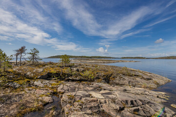 Rocky shoreline with moss and small trees, overlooking a calm, blue lake and distant islands under a bright, clear sky.