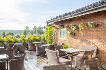 An outdoor patio area with wicker furniture and hanging plants against a brick wall. The patio is situated next to a building with a view of a lush green landscape.