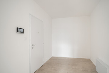 A minimalist entryway with a white door, light wooden flooring, white walls, and a small wall-mounted control panel.