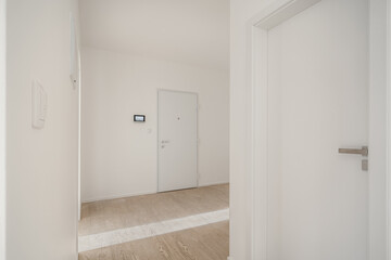 Minimalistic, white hallway with wooden flooring. It features two white doors and a small control panel on the wall.
