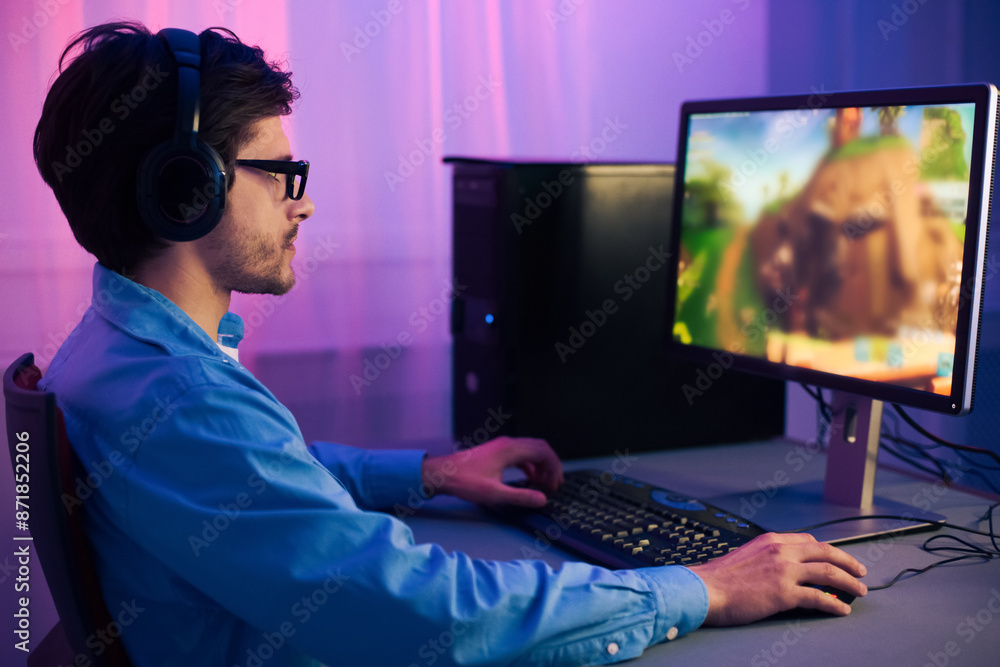 Poster a young man wearing headphones is playing a computer game at a desk in his home office. he is focuse