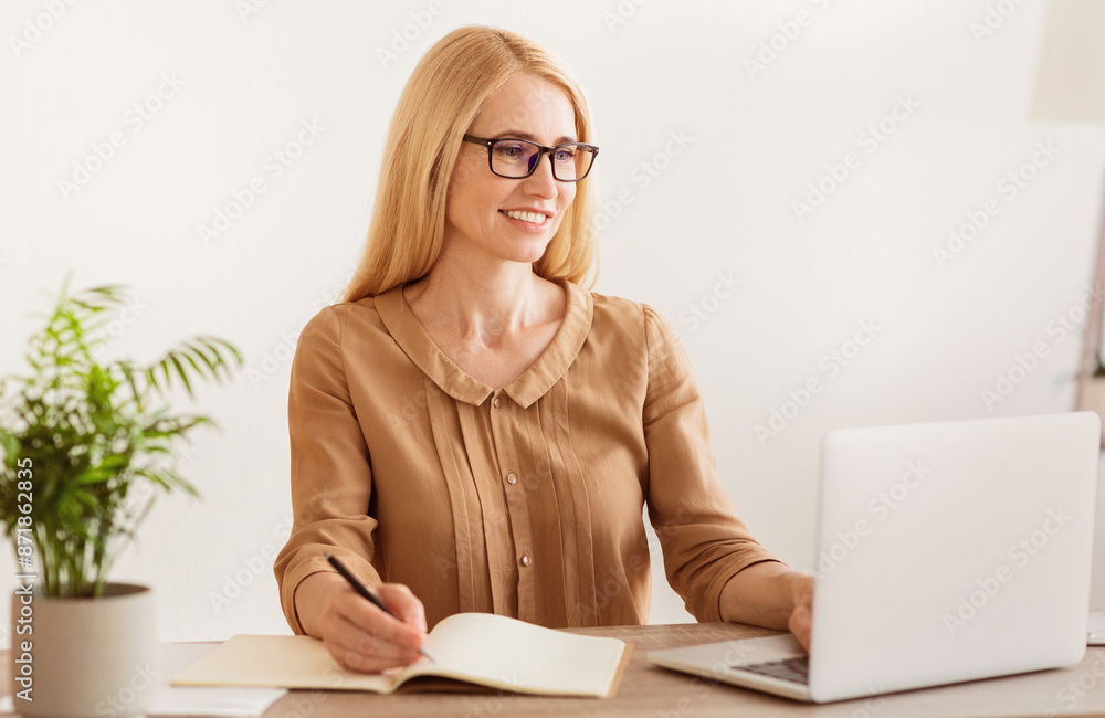 Poster a woman sits at a desk, working on her laptop while taking notes in a notebook. she is smiling and l