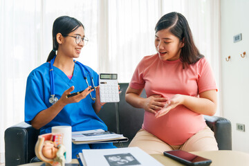 Asian female doctor and nurse consult on a sofa with a pregnant woman, using a calculator to...