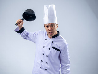 An angry Asian male chef in a white chef's uniform and hat, holding a pan in a raised, aggressive...