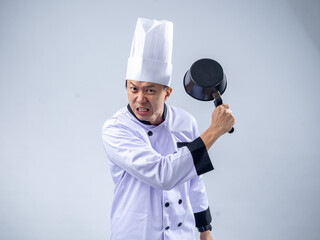 An angry Asian male chef in a white chef's uniform and hat, holding a pan in a raised, aggressive...