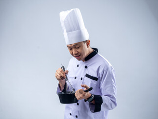 A cheerful Asian male chef in a white chef's uniform and hat, holding a whisk in one hand and a small pan in the other, smiles excitedly against a light blue background