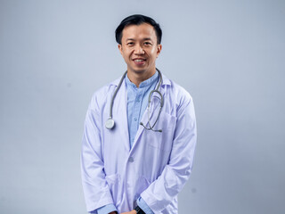 Asian male doctor with a stethoscope around his neck, wearing a white lab coat and light blue shirt, hands in pockets, smiling confidently. Plain background, ideal for healthcare and medical