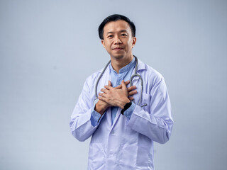 An Asian male doctor in a white coat with a stethoscope, holding his hands over his heart, expresses compassion and care against a light blue background. The setting is professional and reassuring.