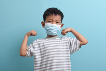 Asian Boy wearing health mask isolated on blue background. Confident kid shows his biceps arm muscle. Medical and health concept