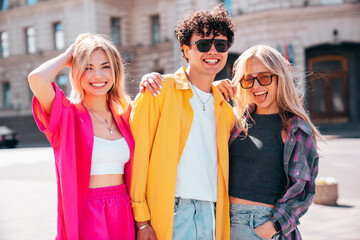 Group of young three stylish friends posing in the street. Fashion man and two cute female dressed in casual summer clothes. Smiling models having fun in sunglasses. Cheerful women and guy outdoors