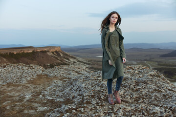 Serene woman standing on hilltop in green coat and boots with hands in pockets overlooking scenic view