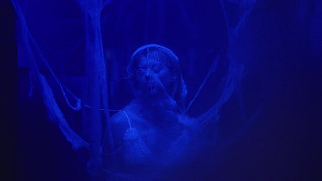 The camera moves through a thick web to a woman covered in web, illuminated by an eerie blue light.
