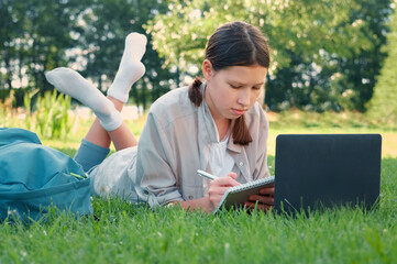 Teenage schoolgirl studying reading her books, tablet and notebook, sitting outdoors. Back to school. Student girl lying on the green grass using laptop in the college yard or park. Distance learning.