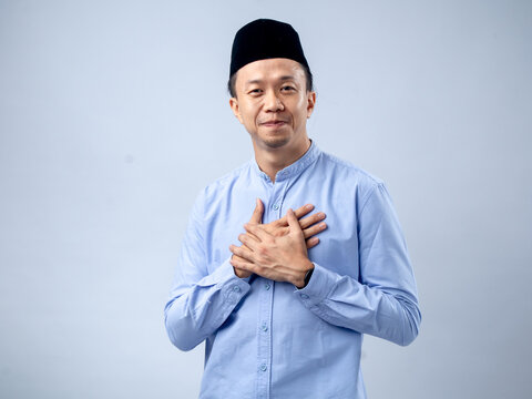 Asian man wearing a black traditional cap and light blue shirt, with hands crossed over his heart, exuding a heartfelt and appreciative expression. The background is plain