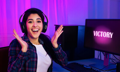 A young woman wearing headphones sits at a computer desk in a dimly lit room, illuminated by pink and blue lights. She is smiling and raising her hands in the air, celebrating her victory.