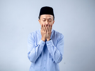 Asian man in traditional black cap and light blue shirt, with hands covering his face in a gesture...