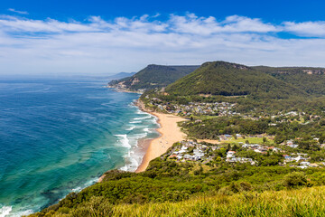 A beautiful aerial view of Clifton and Sea Cliff Bridge with sandy beach and waves crashing on the shore. The beach is bordered by lush green cliffs and the bright blue sky.
