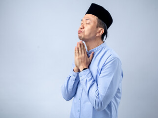 Asian man in traditional black cap and light blue shirt, with hands covering his face in a gesture of prayer or contemplation. The image captures a moment of reflection, set against a plain background