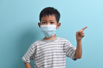Asian Boy wearing health mask, pointing hand to empty background. Isolated on blue background. Medical and health concept. Human population viruses, infections, flu disease prevention