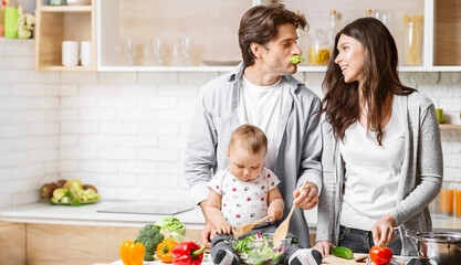 A young couple with their baby are preparing dinner together in a modern kitchen. The father is holding the baby, who is stirring a bowl of salad with a wooden spoon, copy space