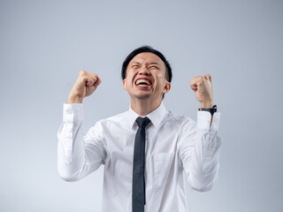 Asian businessman in white shirt and black tie celebrating success with fists clenched and mouth open in excitement, standing against a plain light background. His joyful expression and victory pose