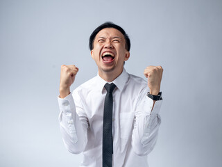Asian businessman in white shirt and black tie celebrating success with fists clenched and mouth open in excitement, standing against a plain light background. His joyful expression and victory pose