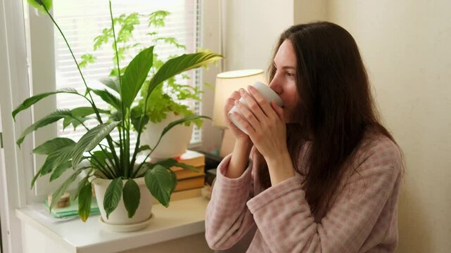 Close-up portrait of an attractive brunette woman holding a cup with her hands and taking a sip of coffee in the kitchen on a sunny morning and looking straight ahead out the window