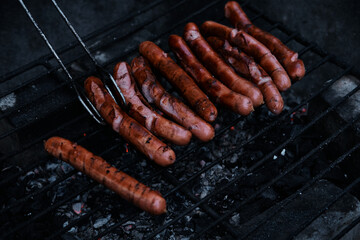 BBQ with fiery sausages on the grill. Grilling food, bbq, barbecue. German sausages are cooked on hot coals in the grill. Close up view, soft blurred background. Flip hot dogs using iron tongs.
