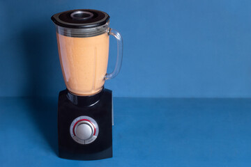 Sleek black blender with a vibrant orange blend of raw ingredients to make gazpacho, set against a blue background. Elegant design with copy space.
